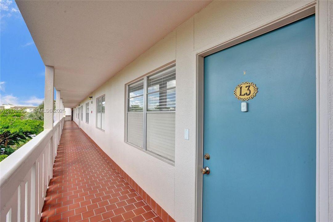 Covered exterior walkway featuring red tile flooring, white railings, and a solid-core door with polished metallic hardware