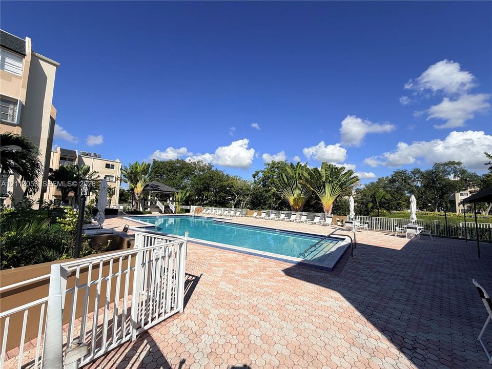 Outdoor swimming pool with a paved deck, white metal perimeter fencing, and mature landscaping
