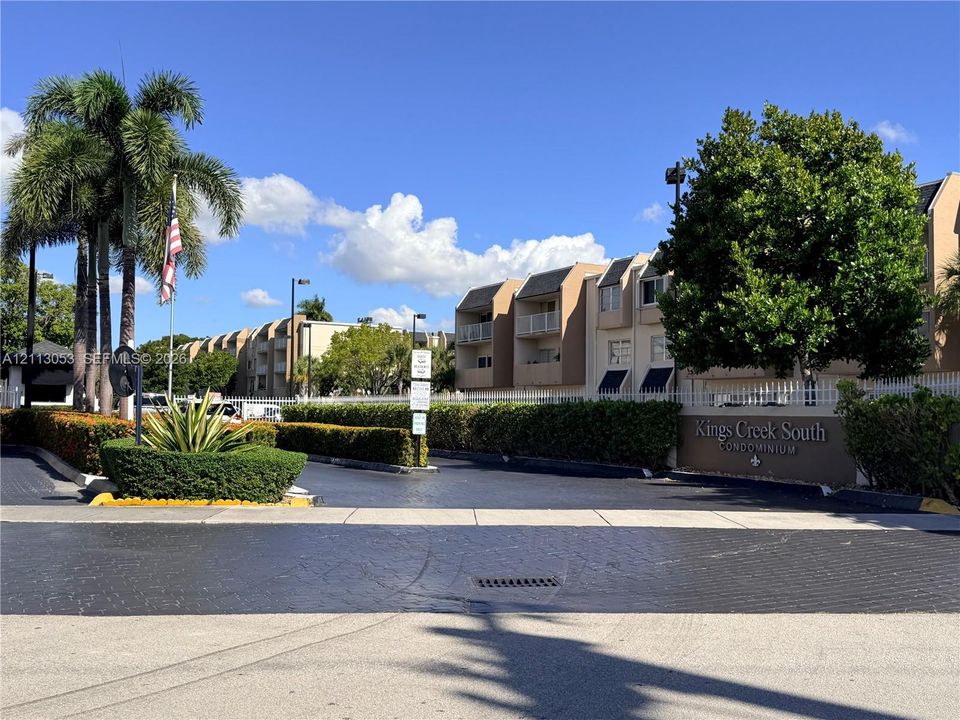 Gated entrance featuring professional landscaping, mature palm trees, multi-story stucco condominium buildings, and private balcony enclosures