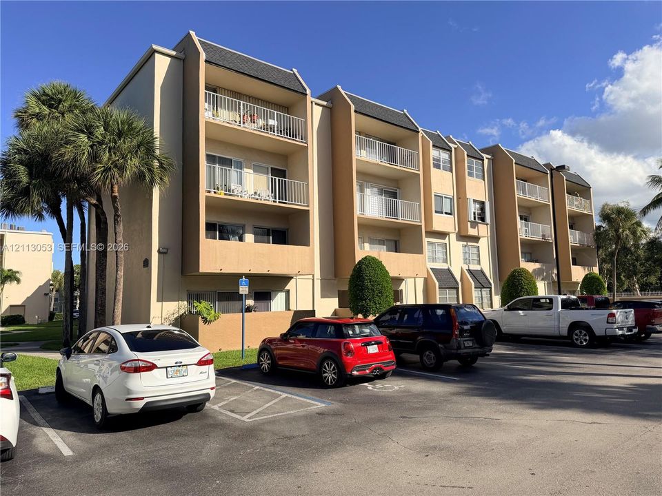 Multi-story exterior featuring a neutral stucco facade, shingled roofing, stacked balconies with metal railings, and mature palm landscaping