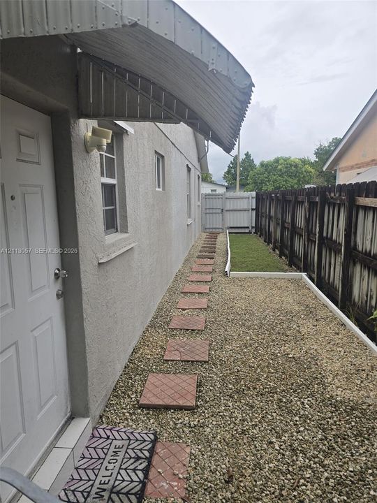 Stucco exterior featuring a metal door awning, stone-paver walkway, gravel landscaping, and a fenced side yard