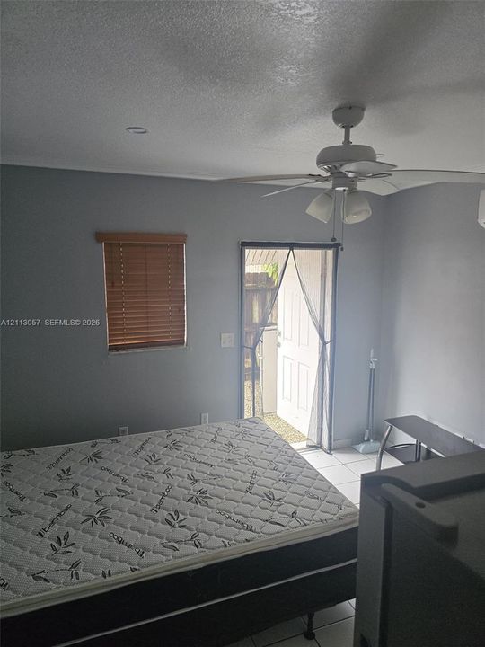 Neutral-toned interior space featuring light-colored tile flooring, a multi-blade ceiling fan with integrated lighting, a single-hung window with wood-finish blinds, and a paneled exterior door