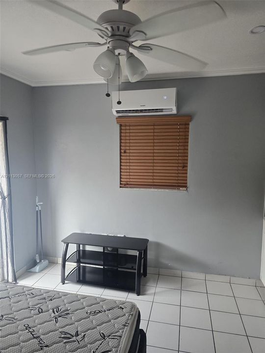 Room featuring white tile flooring, a wall-mounted ductless mini-split air conditioning unit, a ceiling fan with integrated lighting, and horizontal wood-finish blinds