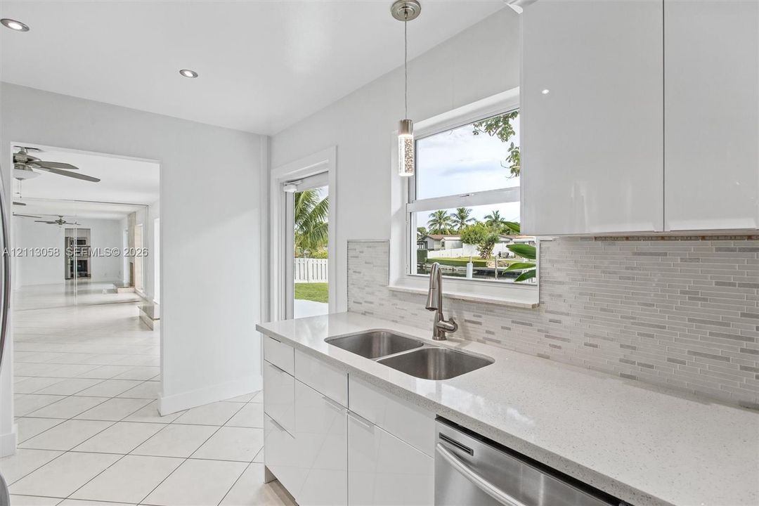 Kitchen featuring high-gloss white cabinetry, quartz countertops, a stone-pattern tile backsplash, stainless steel dual-basin sink with a pull-down faucet, and recessed lighting