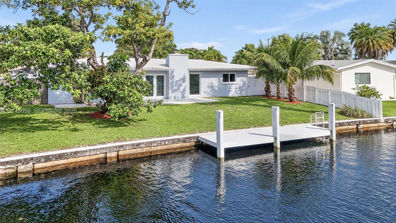 Waterfront residence featuring a private boat dock, concrete seawall, expansive lawn, and white vinyl perimeter fencing