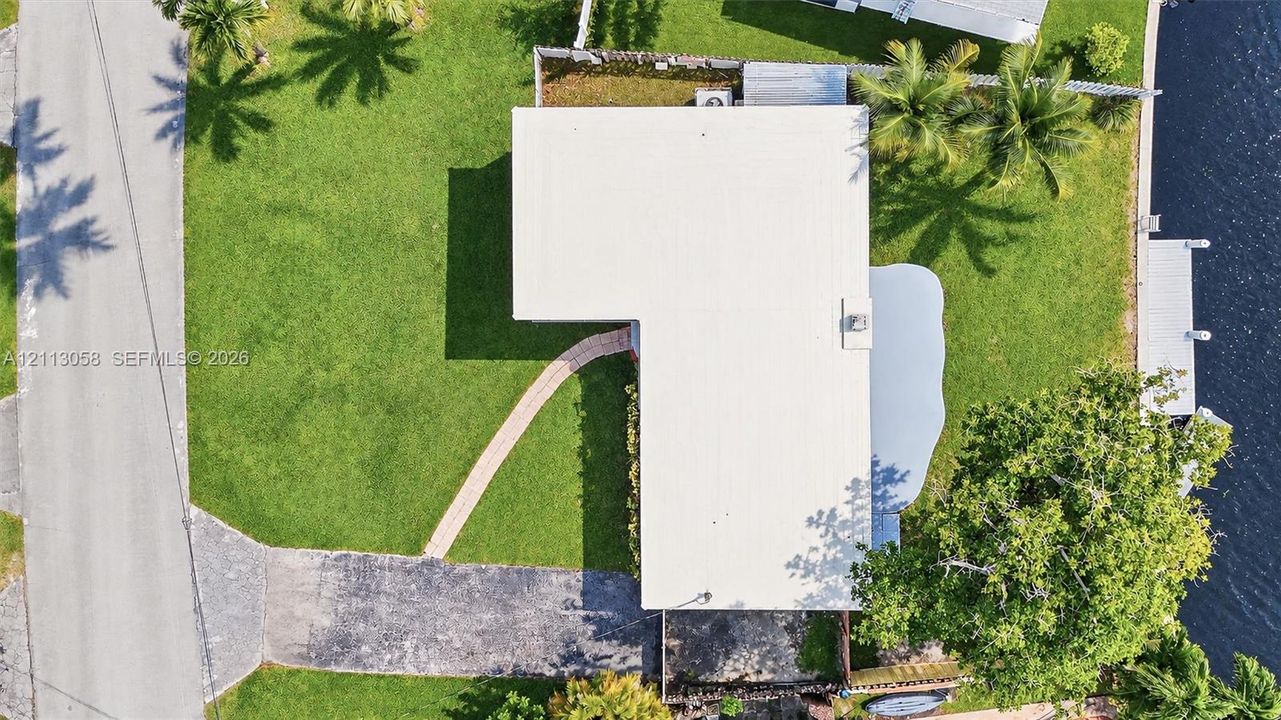 Aerial perspective of a waterfront lot featuring a private dock, asphalt driveway, meandering paved walkway, and landscaped lawn with mature trees