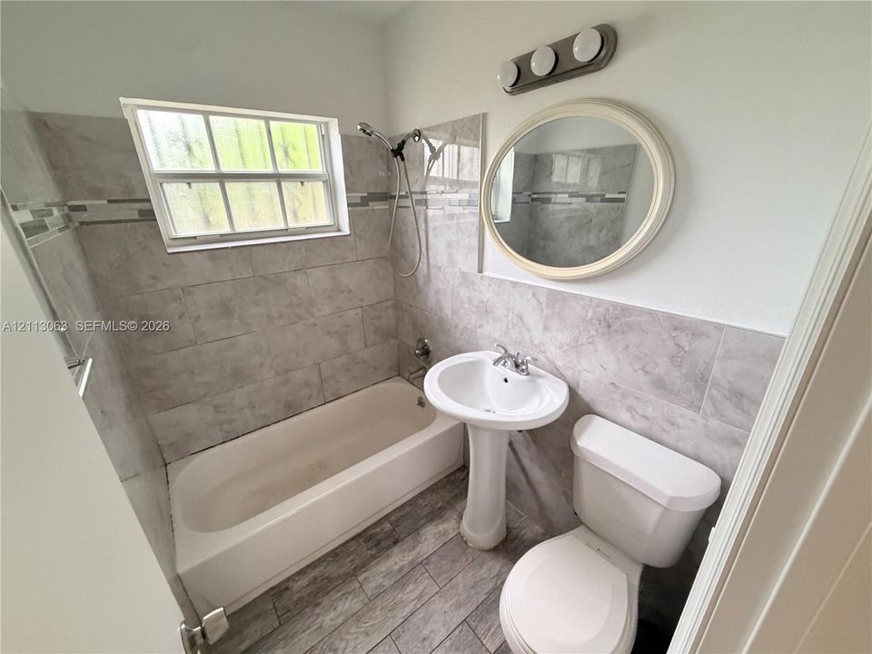 Bathroom featuring tile-surround bathtub with shower head, pedestal sink, oval wall-mounted mirror, and wood-finish flooring