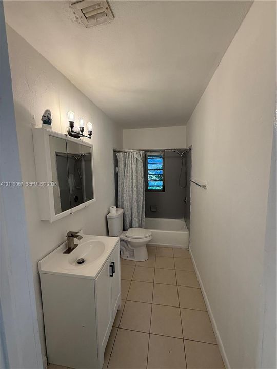 Bathroom featuring a white vanity cabinet with a integrated sink, three-light wall-mounted fixture, mirrored medicine cabinet, neutral floor tiling, and a tub-shower combination with a louvered window