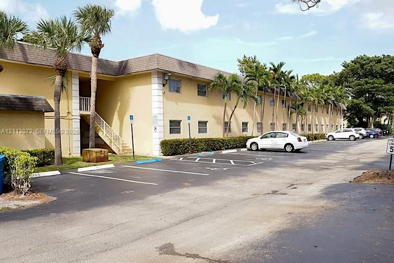 Two-story exterior featuring light-toned masonry walls, defined vertical quoin detailing, a shingled roof, mature palm tree landscaping, and paved surface parking