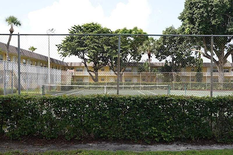 Fenced tennis court with green surfacing, surrounded by mature trees and manicured hedges