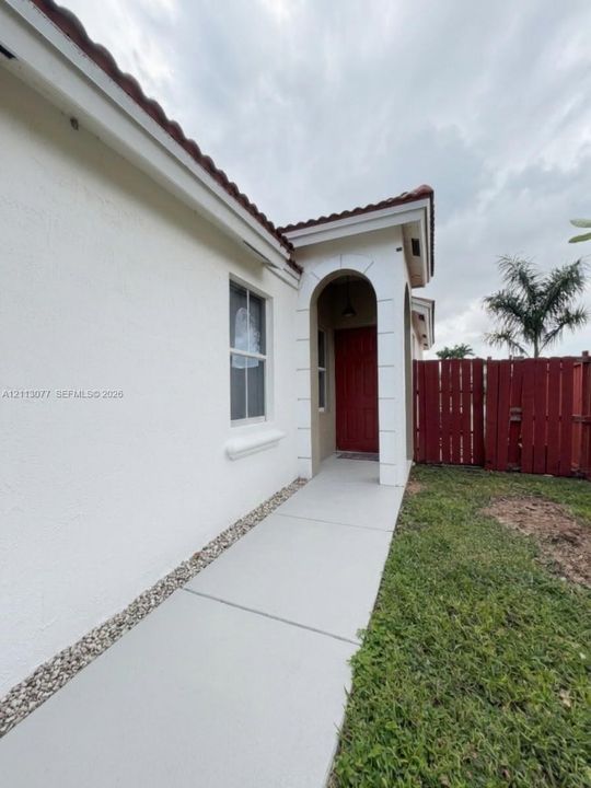 Stucco exterior featuring a covered entryway with a recessed arch, red paneled door, concrete walkway, and wood-slat perimeter fencing