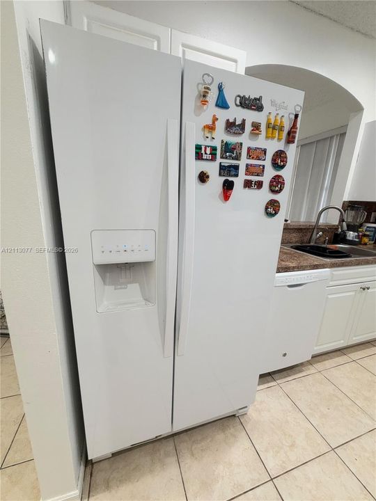 White side-by-side refrigerator with exterior water and ice dispenser, complemented by white cabinetry, solid-surface countertops, a stainless steel sink with a high-arc faucet, and tiled flooring