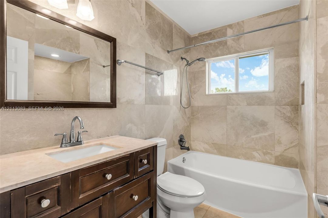 Bathroom featuring floor-to-ceiling large-format tile walls, dark wood cabinetry with a neutral stone countertop, undermount sink with a gooseneck faucet, and a tub-shower combination with a detachable showerhead