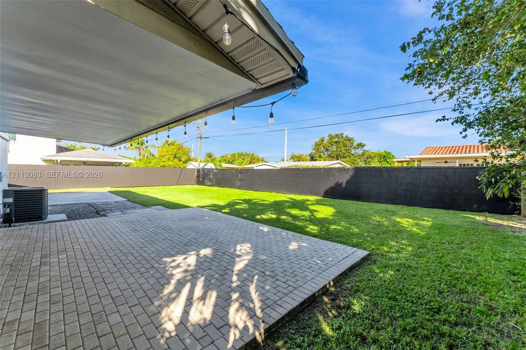 Backyard featuring a paved brick patio, expansive grass lawn, solid perimeter fencing, and an overhead structure with attached string lighting