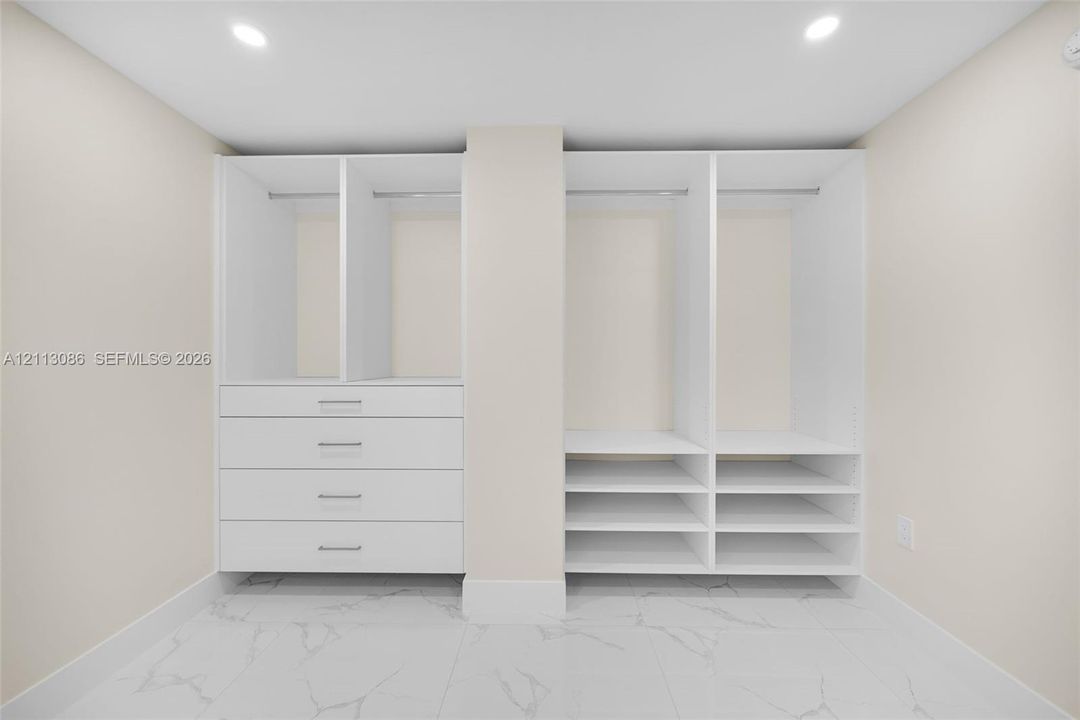 Custom white built-in cabinetry featuring hanging rods, four integrated storage drawers, and multiple open shelving units set against large-format, light-toned stone-finish flooring with prominent veining