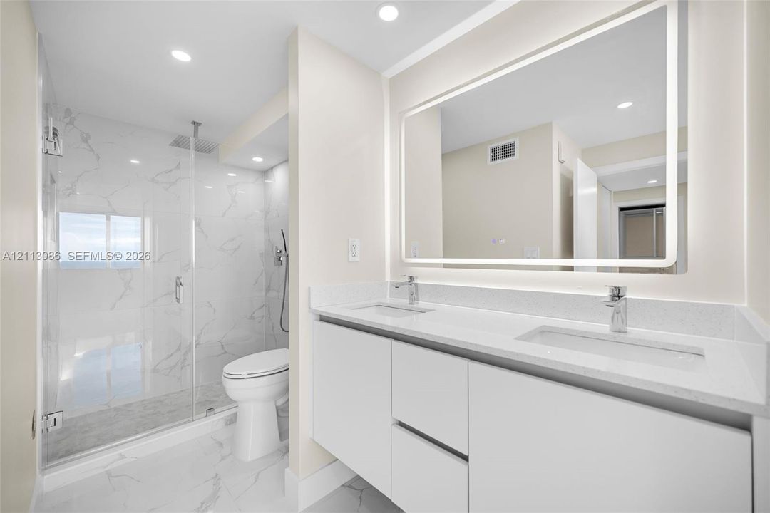 Dual-sink vanity with white cabinetry and solid surface countertop, backlit vanity mirror, walk-in glass shower enclosure featuring rainfall shower head and floor-to-ceiling stone-finish tile surround, and coordinating stone-finish flooring