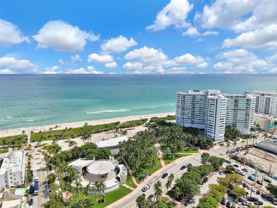 High-altitude coastal setting featuring a sandy shoreline, turquoise waters, integrated park space with mature palm trees, and an open-air pavilion