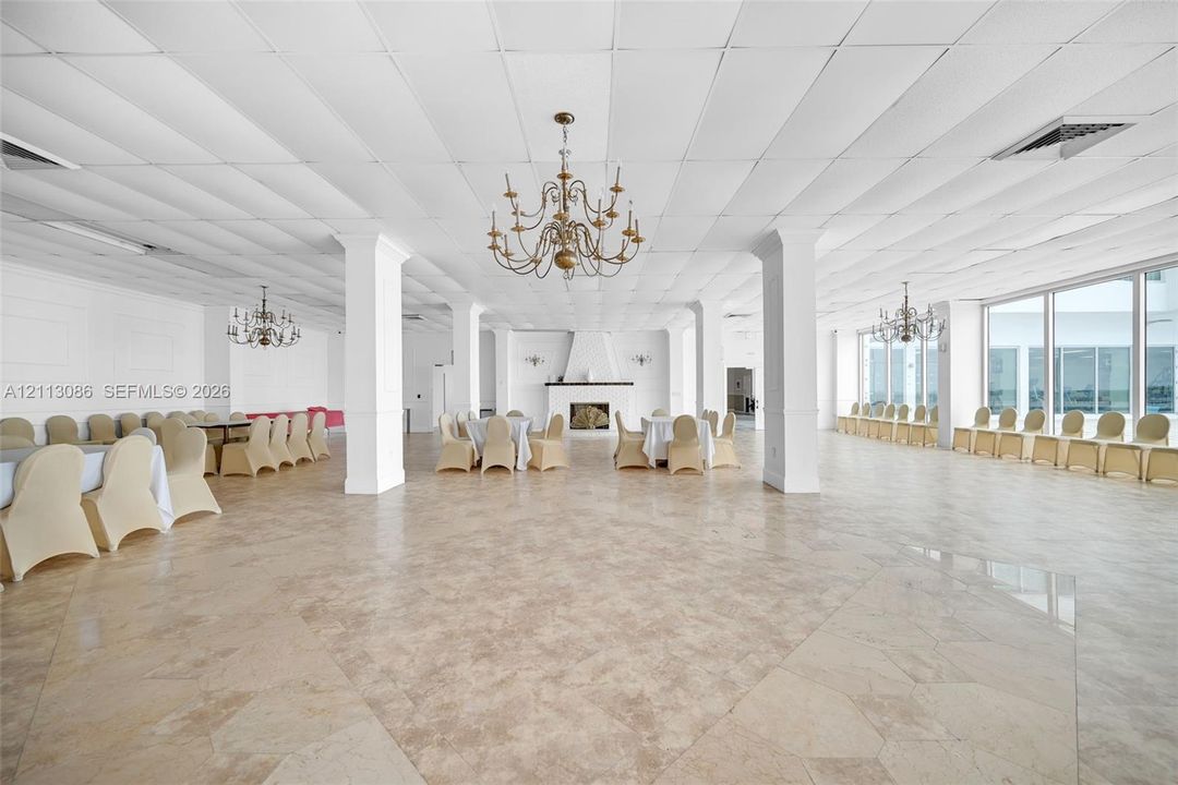 Expansive open-concept hall featuring large-format tile flooring, a central fireplace with a decorative mantel, structural columns, ornate multi-arm chandeliers, and floor-to-ceiling windows