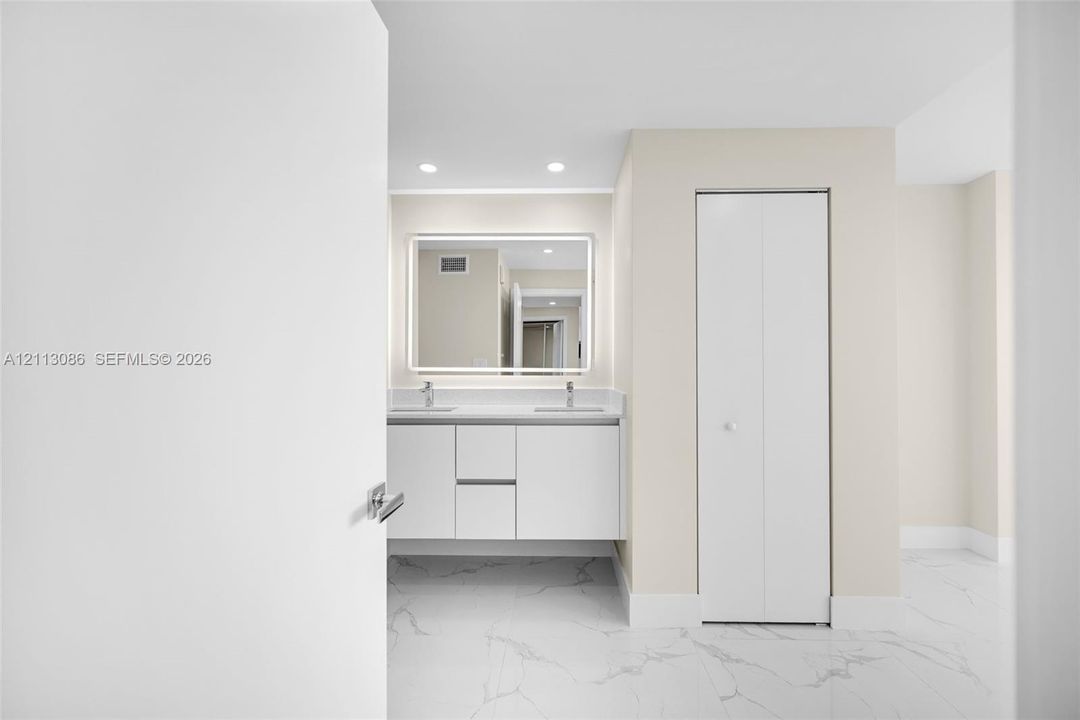 Floating vanity with double under-mount sinks, integrated stone-surface countertop, LED-backlit mirror, and white bi-fold closet doors, finished with large-format light-toned stone-patterned flooring