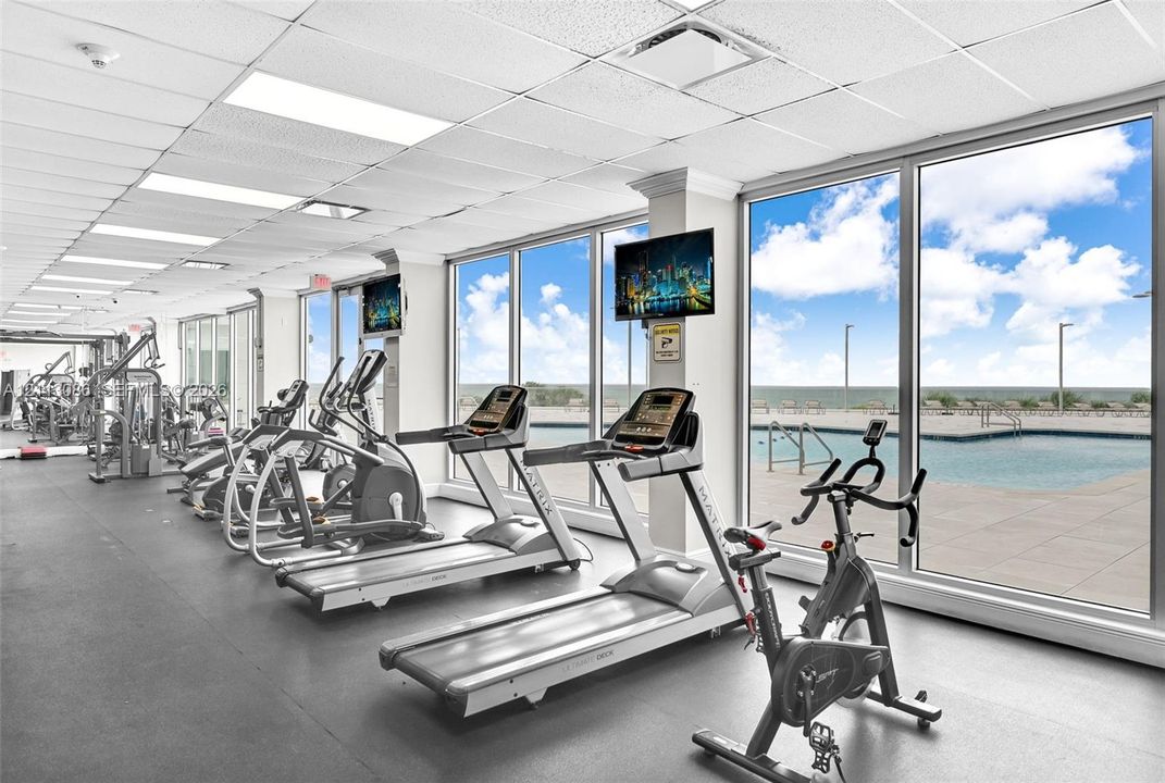 Fitness center featuring wall-to-wall windows, expansive natural light, rubber flooring, drop ceiling panels, and direct sightlines to an outdoor pool