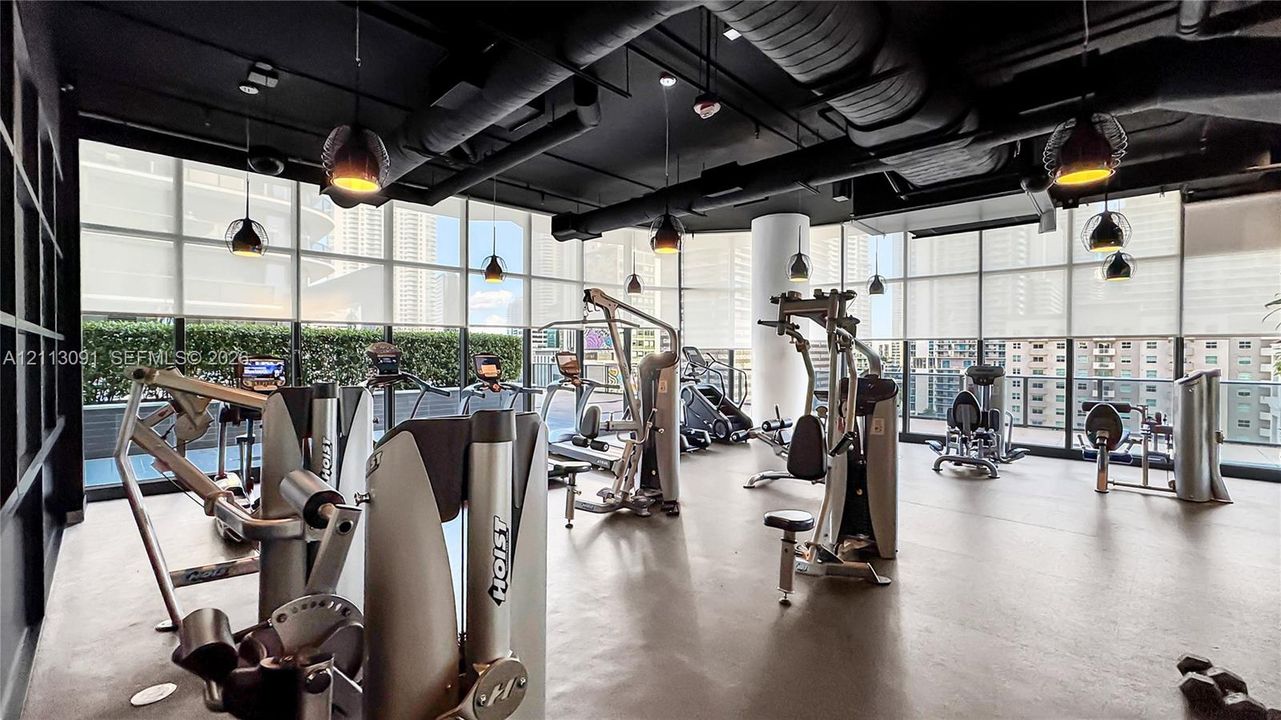 Fitness center featuring floor-to-ceiling windows, exposed industrial ductwork, black ceilings, and pendant lighting