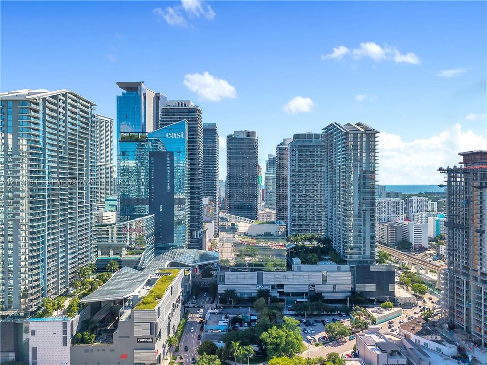 Aerial perspective of a high-density urban corridor featuring glass-facade skyscrapers, multi-level retail complexes, elevated transit infrastructure, and manicured green rooftops