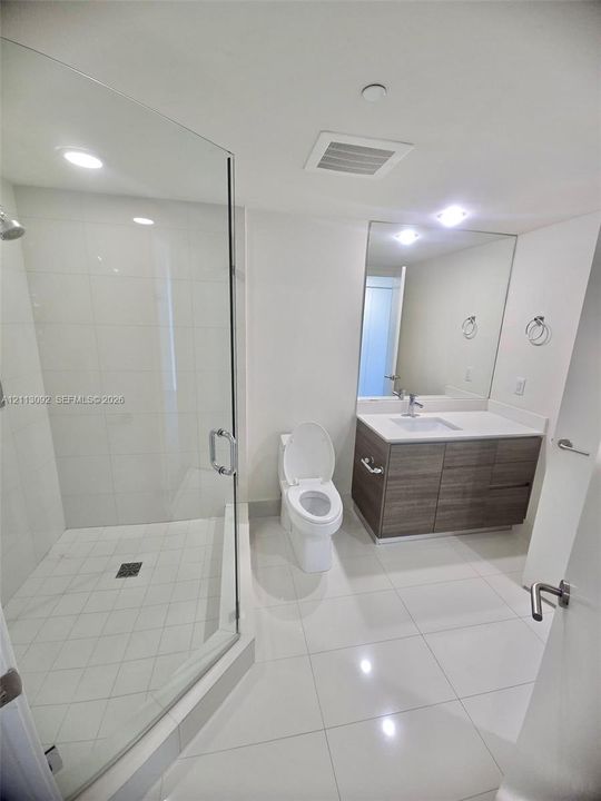 Glass-enclosed shower with tiled surround, wood-finish vanity with stone countertop, large wall-mounted mirror, and polished tile flooring