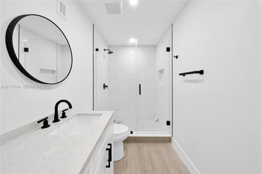 Bathroom featuring a white vanity with a neutral stone-look countertop, circular black-framed mirror, wood-finish flooring, and a walk-in shower with a glass enclosure and matte black fixtures