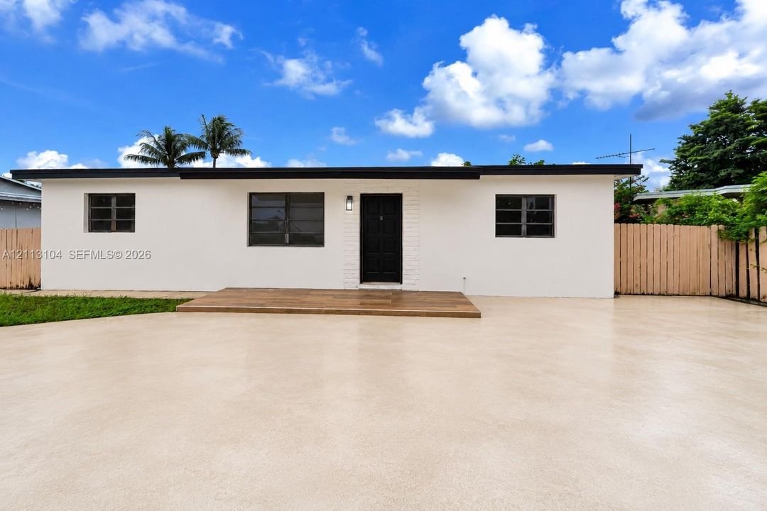 Single-story residence featuring a smooth stucco exterior, dark-toned metal-framed windows, a wood-plank entryway deck, and an expansive concrete paved area
