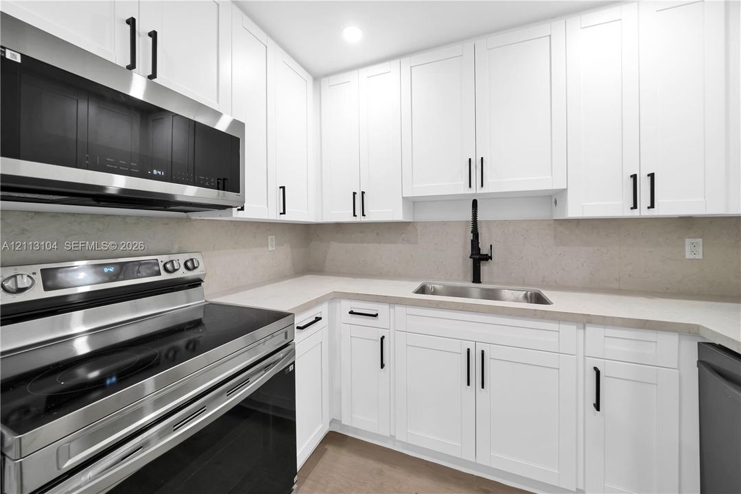 Kitchen featuring white shaker-style cabinetry, stainless steel appliances, neutral stone-finish countertops, matte black hardware, and an undermount sink with a high-arc matte black faucet