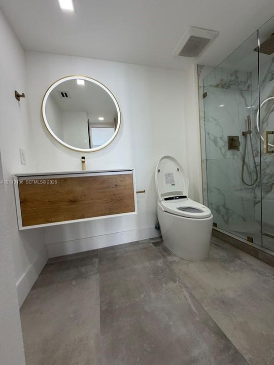Floating vanity with wood-finish cabinetry, round backlit mirror, large-format floor tile, and glass-enclosed shower with stone-surround wall detailing