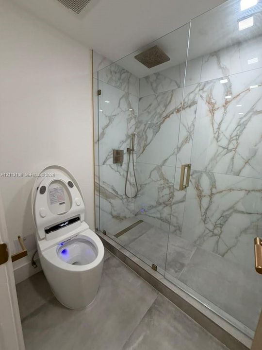 Walk-in shower featuring glass paneling, gold-tone fixtures, rain-style showerhead, and large-format wall tile with a veined pattern, complemented by a smart toilet with integrated lighting