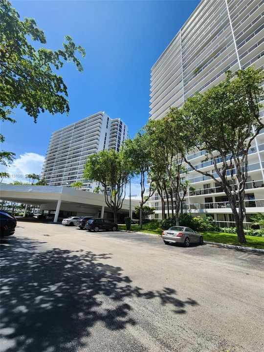 High-rise residential exterior featuring white concrete facade, wrap-around balcony railings, covered arrival area, and mature landscaping