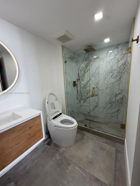 Floating wood-finish vanity with an integrated basin, frameless glass walk-in shower with stone-patterned tile surround, brass-finish shower hardware, and high-efficiency smart toilet