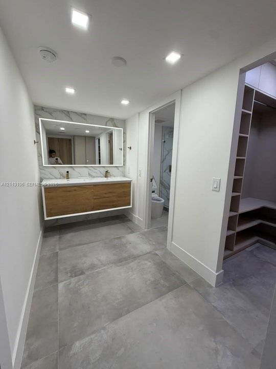 Floating wood-finish vanity with dual sinks, backlit wall-mounted mirror, large-format tile flooring, and built-in walk-in closet cabinetry