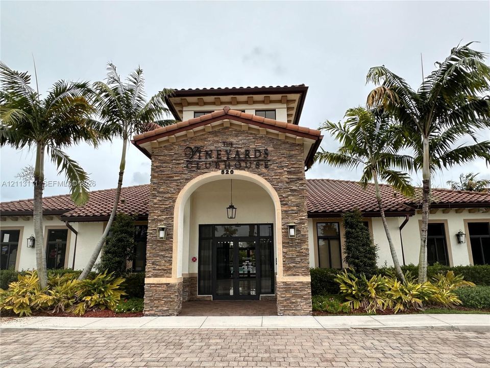 Clubhouse exterior featuring a stone-clad facade, arched entryway, terracotta tile roof, and brick-paver driveway