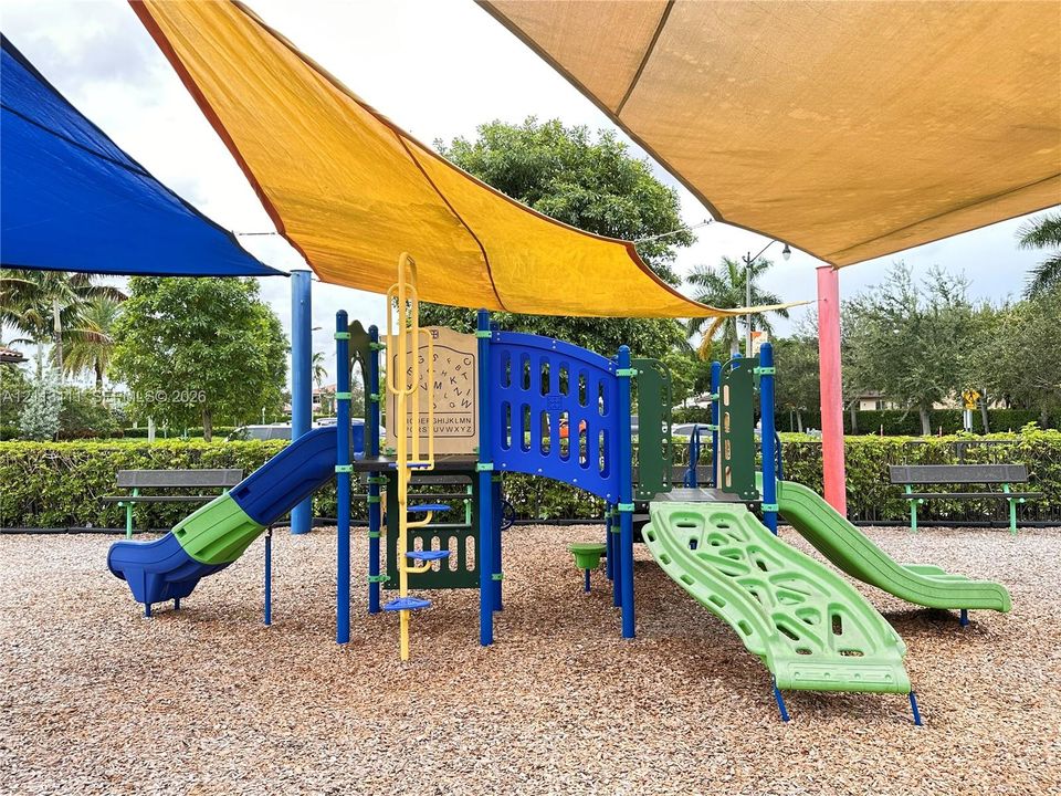 Outdoor playground featuring multi-colored slides, climbing structures, and overhead sun shades, set within a wood-mulched area with peripheral metal seating
