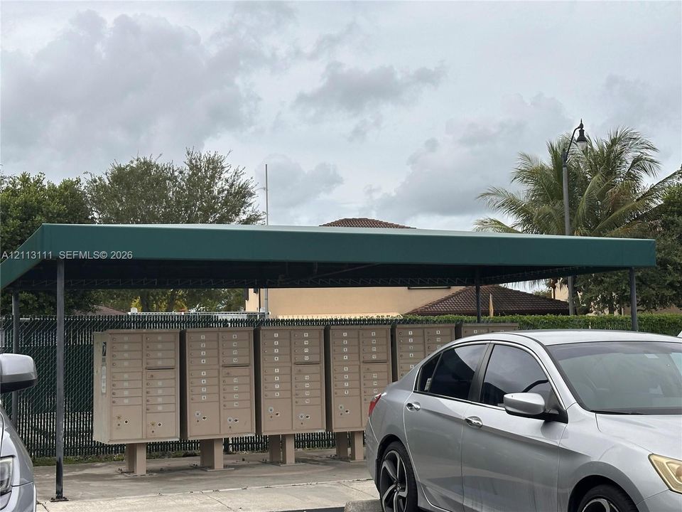 Covered communal mailbox station featuring a dark green metal awning, concrete base, and multi-unit locking mailboxes