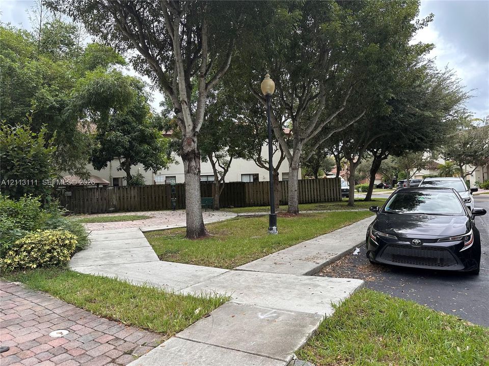 Landscaped green space featuring mature trees, concrete walkways, a paver stone patio area, and a decorative street-style lamp post