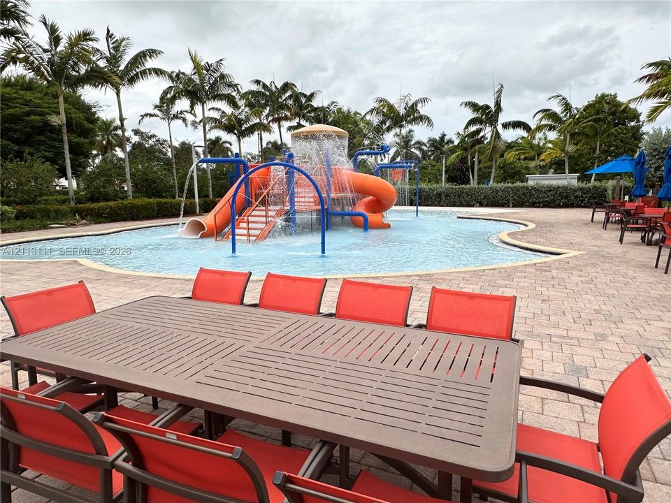Resort-style pool featuring a splash pad with water slides and built-in water features, surrounded by a paved patio deck and mature palm trees