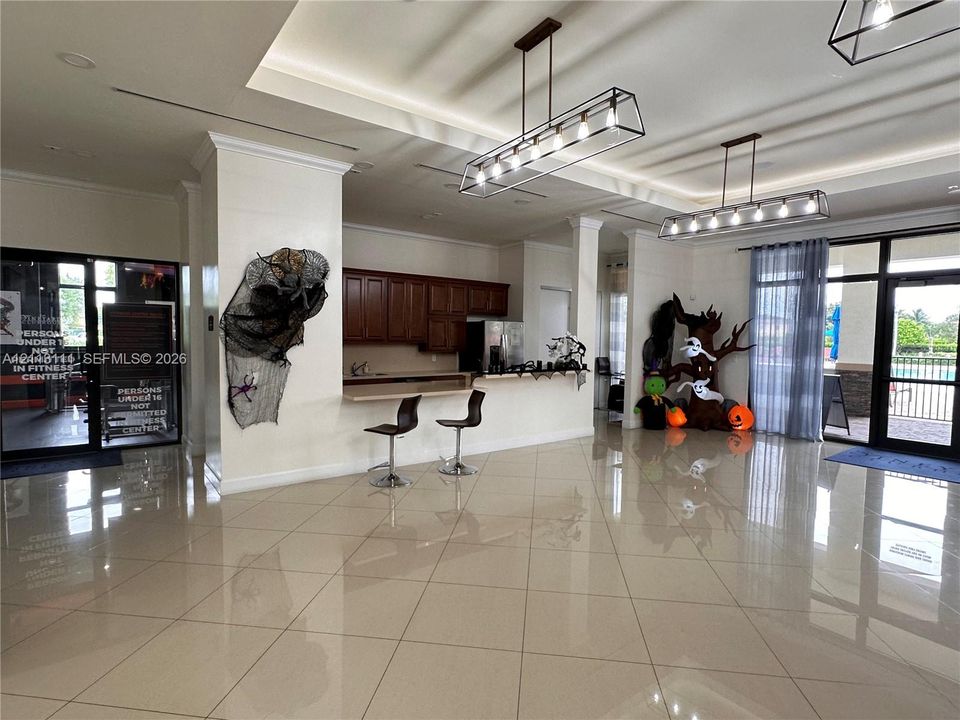Large communal area featuring oversized tile flooring, recessed tray ceiling, dual linear metal pendant light fixtures, and a wet bar with wood-finish cabinetry and a service counter