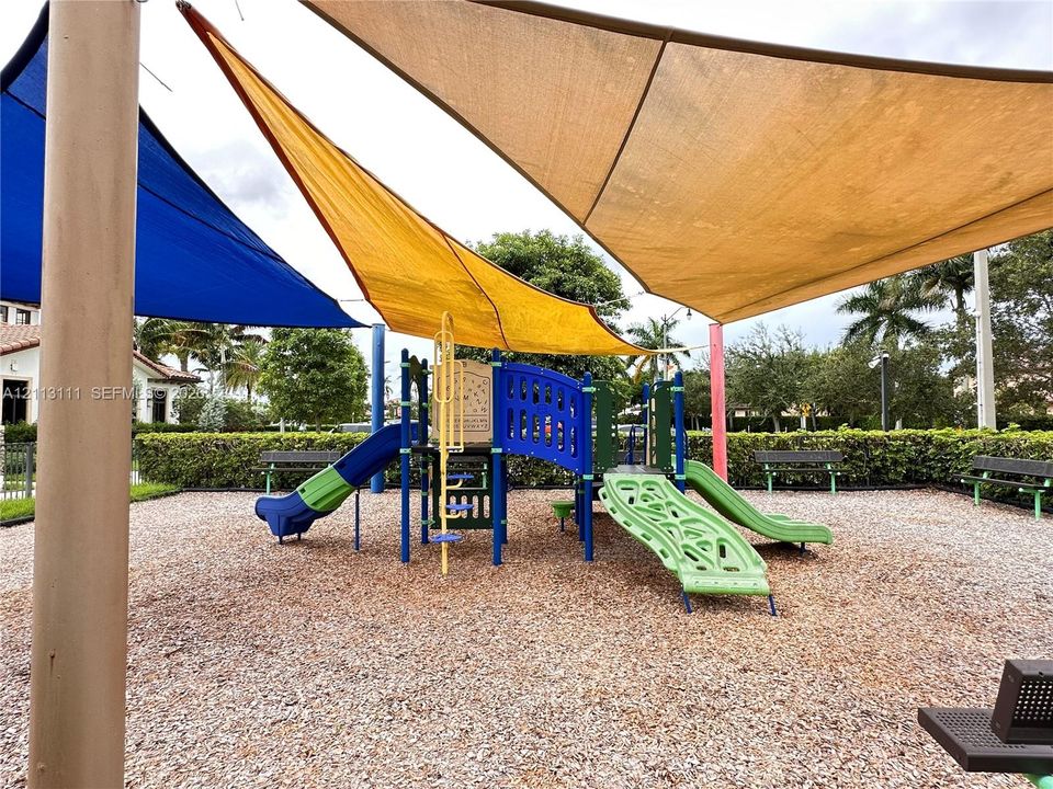 Outdoor recreational area featuring multiple shade sails, a multi-component play structure with dual slides, mulch ground cover, and metal bench seating
