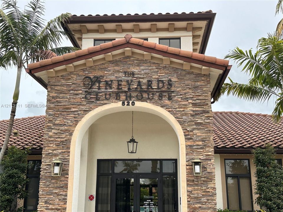 Clubhouse exterior featuring stacked stone masonry, a covered arched entryway, clay tile roofing, decorative wooden corbels, and metal exterior light fixtures