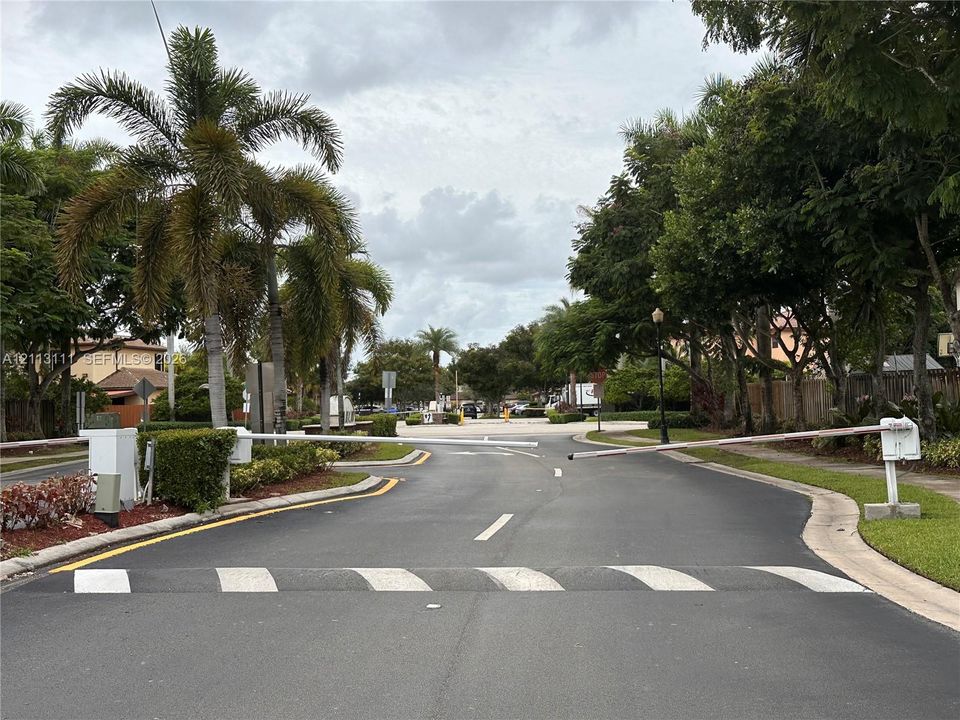 Gated community entrance featuring motorized security arms, asphalt roadway, concrete speed bumps, and mature palm and hardwood landscaping