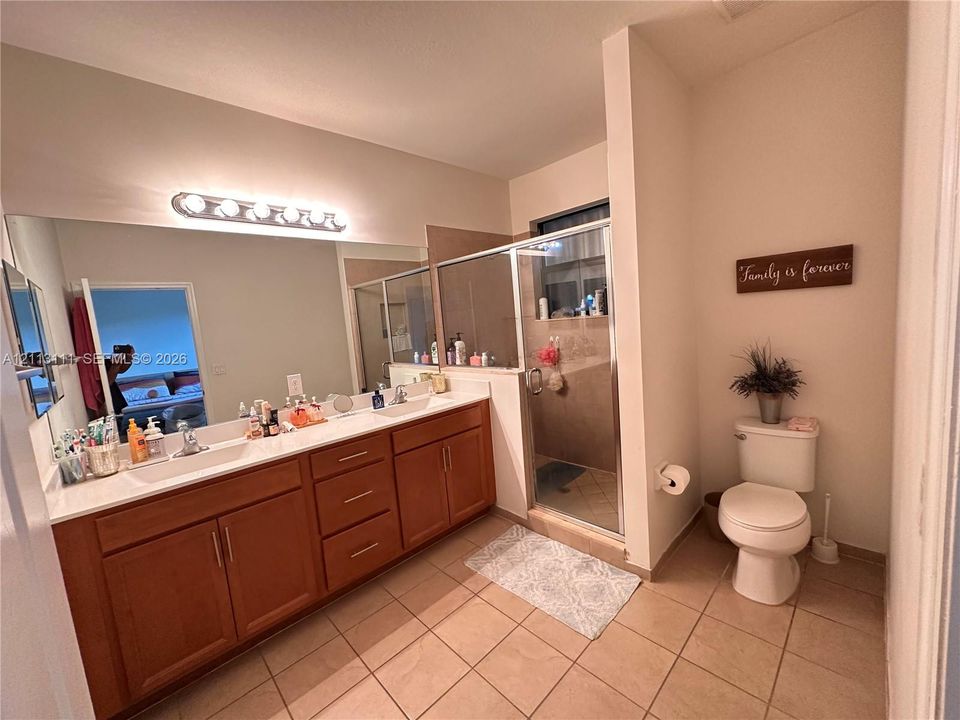 Bathroom featuring a dual-sink vanity with wood cabinetry, solid surface countertop, large wall-mounted mirror, and a glass-enclosed shower with tile surround