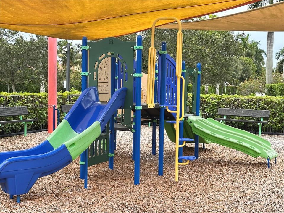 Outdoor recreation area featuring a multi-level playground structure with dual slides, protective overhead shade canopy, wood chip ground cover, and surrounding park benches