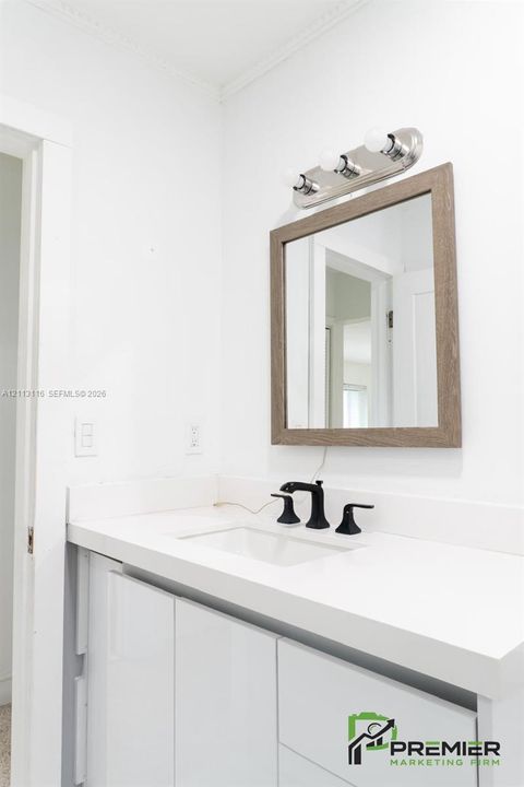Glossy white vanity featuring a solid surface countertop, undermount sink, matte black widespread faucet, wood-toned framed mirror, and a three-light vanity fixture