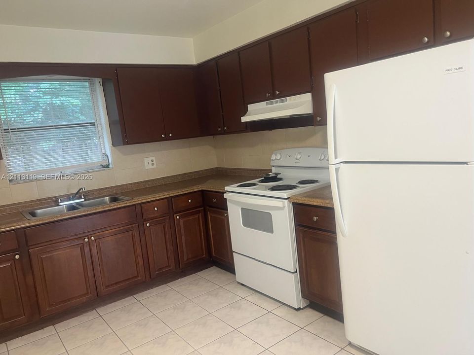 Kitchen featuring dark-toned wooden cabinetry, tiled flooring, laminate countertops, a double-basin stainless steel sink, and white appliances