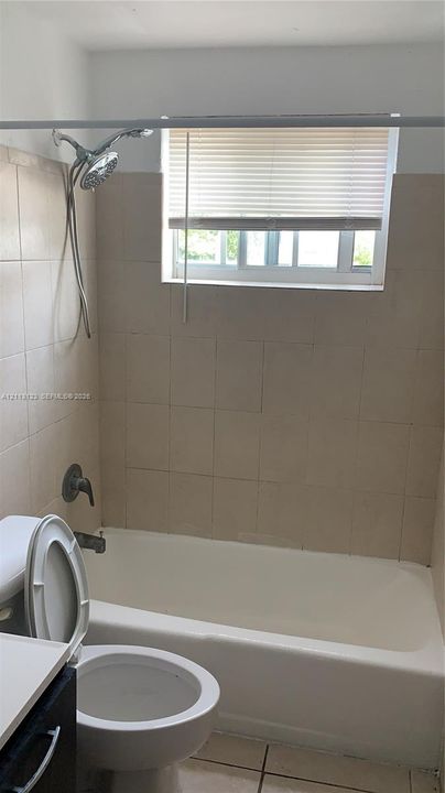 Full-size bathtub with surrounding wall tile, handheld shower fixture, and window with built-in blinds