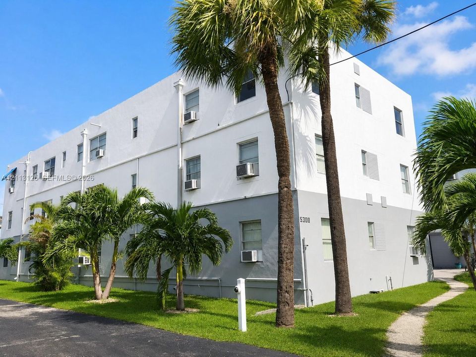 Multi-story exterior featuring a two-tone white and grey facade, mature palm landscaping, and a gravel walkway